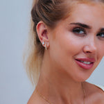 A close-up of a woman's ear wearing small gold earrings, including a huggie hoop and a stud with a clear stone, against a light gray background.
