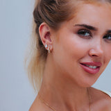 A close-up of a woman's ear wearing small gold earrings, including a huggie hoop and a stud with a clear stone, against a light gray background.