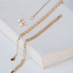 Gold chain jewelry set with a tennis-style chain, a delicate chain with a small charm, and a pair of stud earrings with clear stones, arranged on an open book.