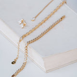 Gold chain jewelry set with a tennis-style chain, a delicate chain with a small charm, and a pair of stud earrings with clear stones, arranged on an open book.