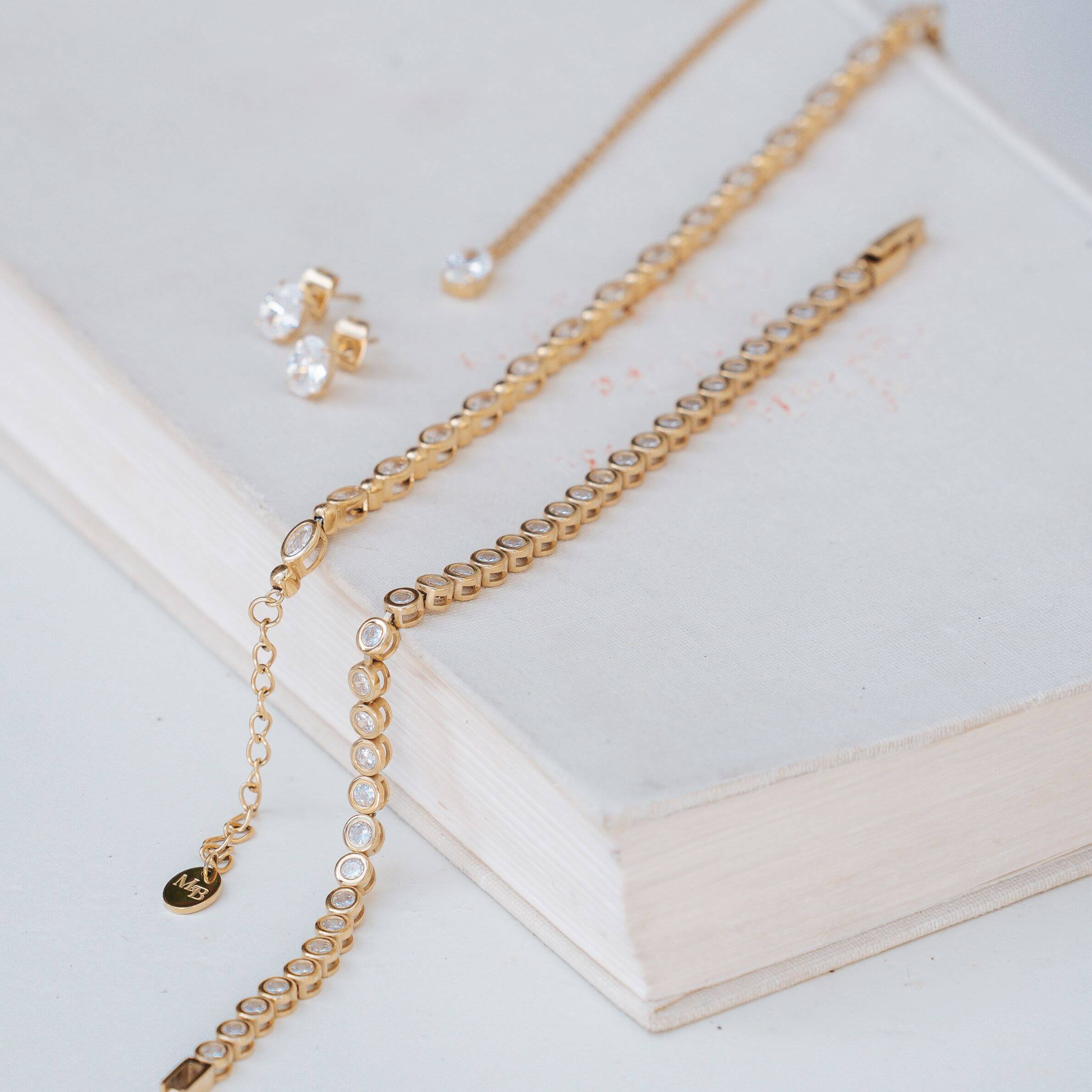 Gold chain jewelry set with a tennis-style chain, a delicate chain with a small charm, and a pair of stud earrings with clear stones, arranged on an open book.