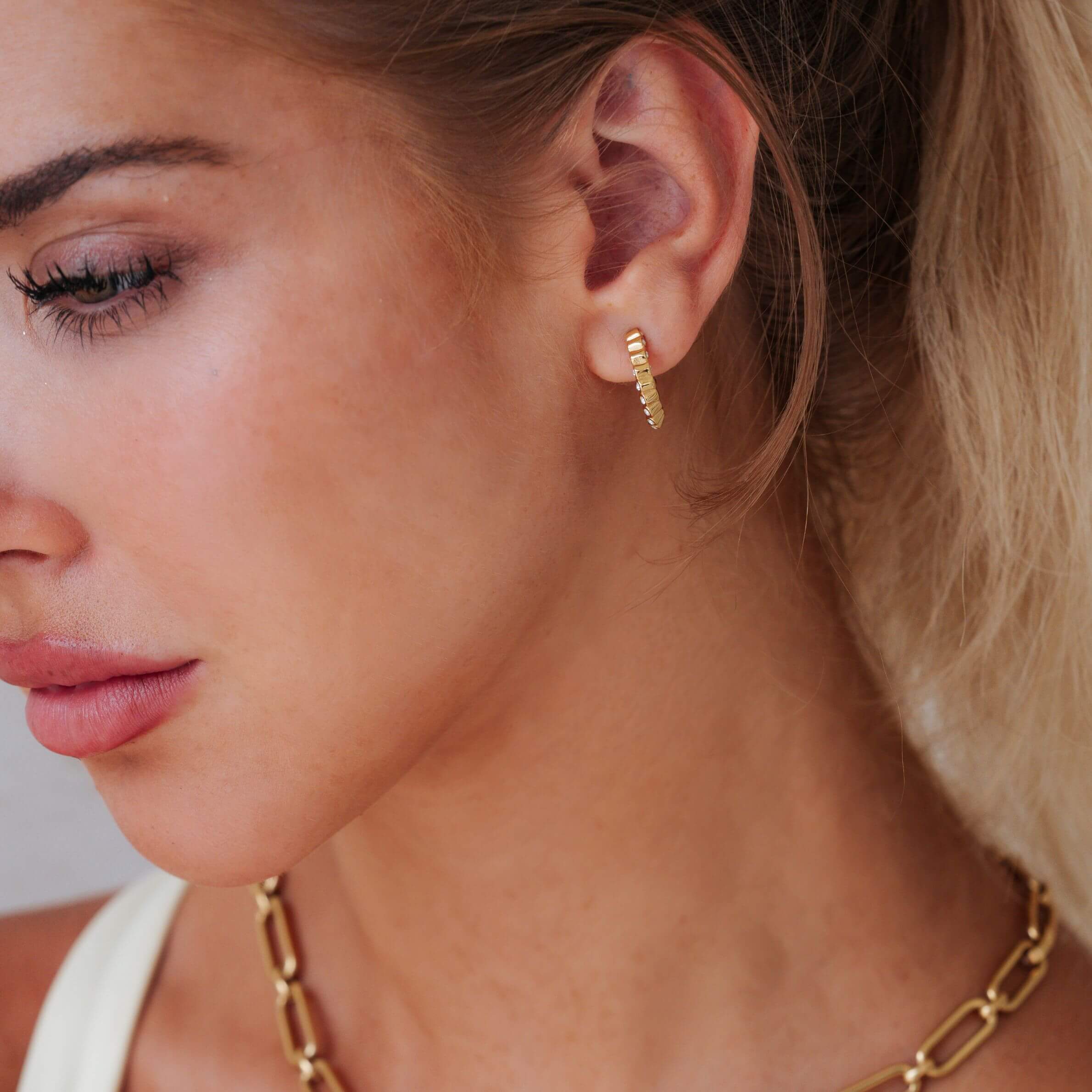 A close-up of a woman's ear wearing a small gold hoop earring, with her hair pulled back and a gold chain necklace partially visible.