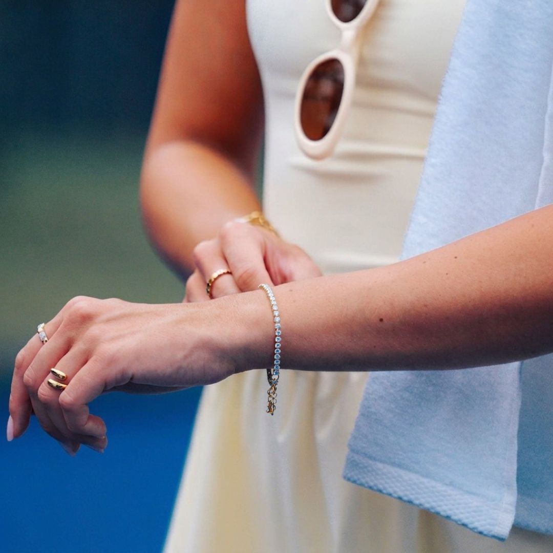 A close-up lifestyle shot of a model wearing an elegant gold tennis bracelet on her wrist, alongside other gold rings, a yellow-coloured tennis dress, and a white towel. She is on a tennis court. 