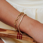 A close-up of a person's wrist wearing two delicate bracelets: one gold chain with elongated links and one gold chain with small clear stones, resting on a wooden surface.