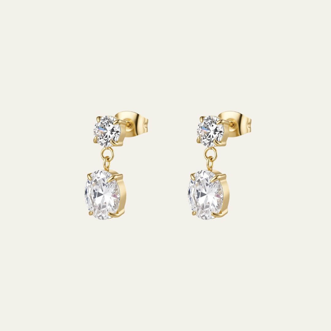 A pair of elegant gold drop earrings with a small round clear gemstone above a larger oval clear gemstone, shown on a light cream background. They were designed for bridal by Mettle & Bloom. 