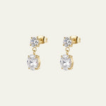A pair of elegant gold drop earrings with a small round clear gemstone above a larger oval clear gemstone, shown on a light cream background. They were designed for bridal by Mettle & Bloom. 