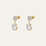 A pair of elegant gold drop earrings with a small round clear gemstone above a larger oval clear gemstone, shown on a light cream background. They were designed for bridal by Mettle & Bloom. 