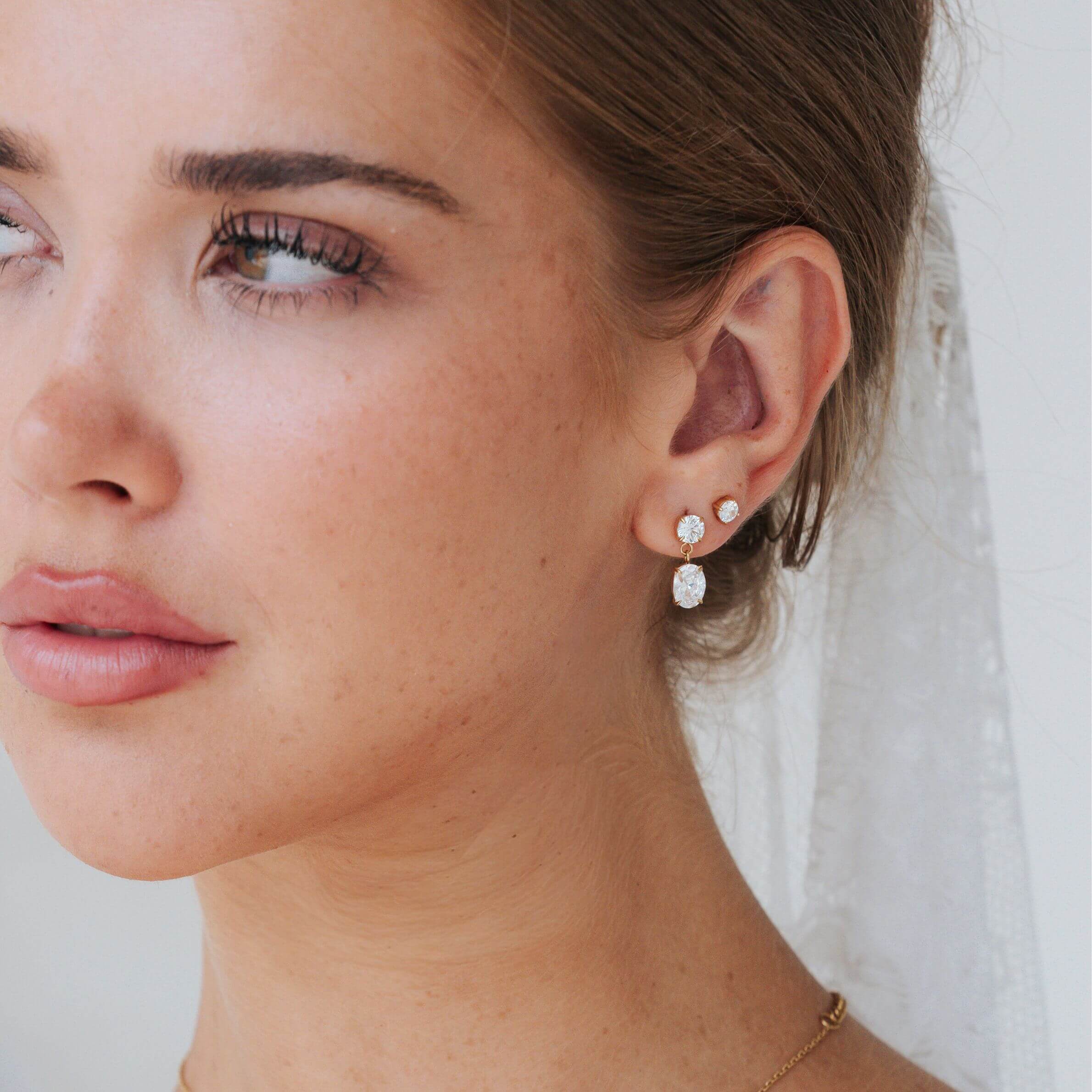 A close-up of a woman's ear wearing a pair of small gold drop earrings with clear teardrop-shaped stones and round clear stones, against a soft white bridal veil background.