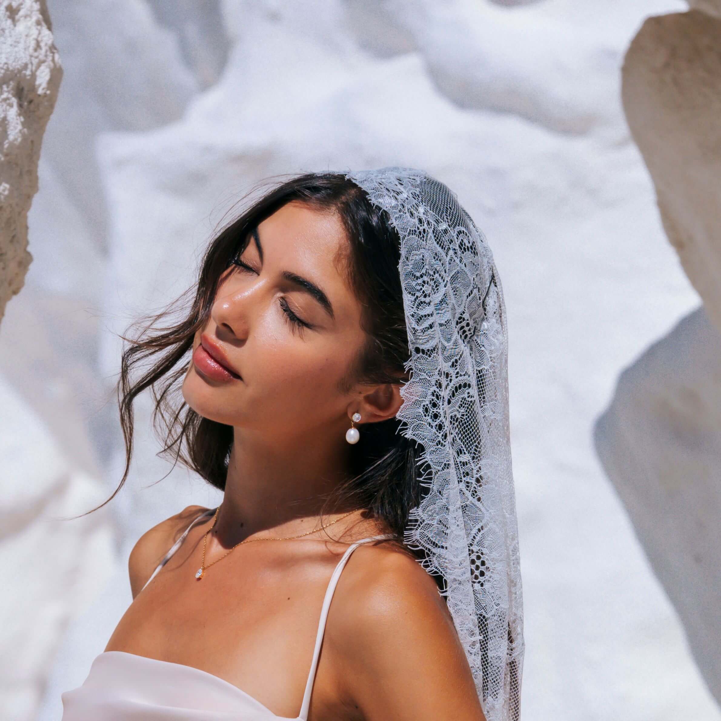 A bride outdoors wearing a white lace bridal veil and a white spaghetti-strap dress, with rocky white surroundings and bright sunlight. She has a pair of Heirloom earrings from Mettle & Bloom on. The stud is a white dazzling stone, suspended from it is a larger pearl. 