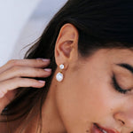 A close-up lifestyle shot of a woman wearing a gold drop earring with a white pearl and a clear crystal accent. The earring is shown on her earlobe, with her hand resting near her hair.