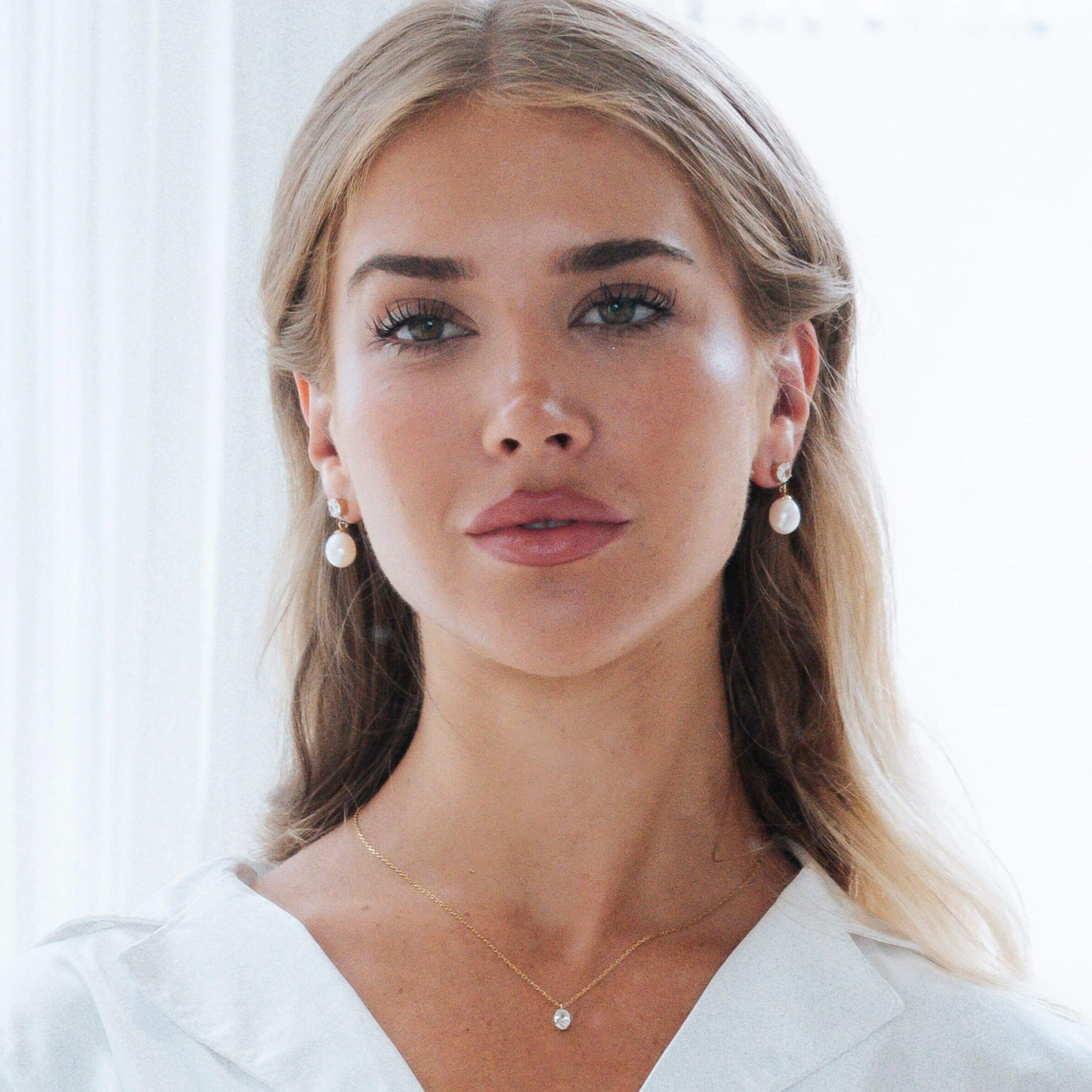 A close-up portrait of a woman with blonde hair wearing matching pearl drop earrings and a delicate gold necklace with a small clear stone, against a bright white background.