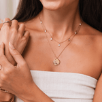 Woman wearing a gold necklace with a hexagonal pendant, holding her arm.