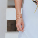 A close-up of a person's hand and wrist wearing two delicate gold bracelets, one beaded and one slim with a small clear stone, while wearing a white dress.