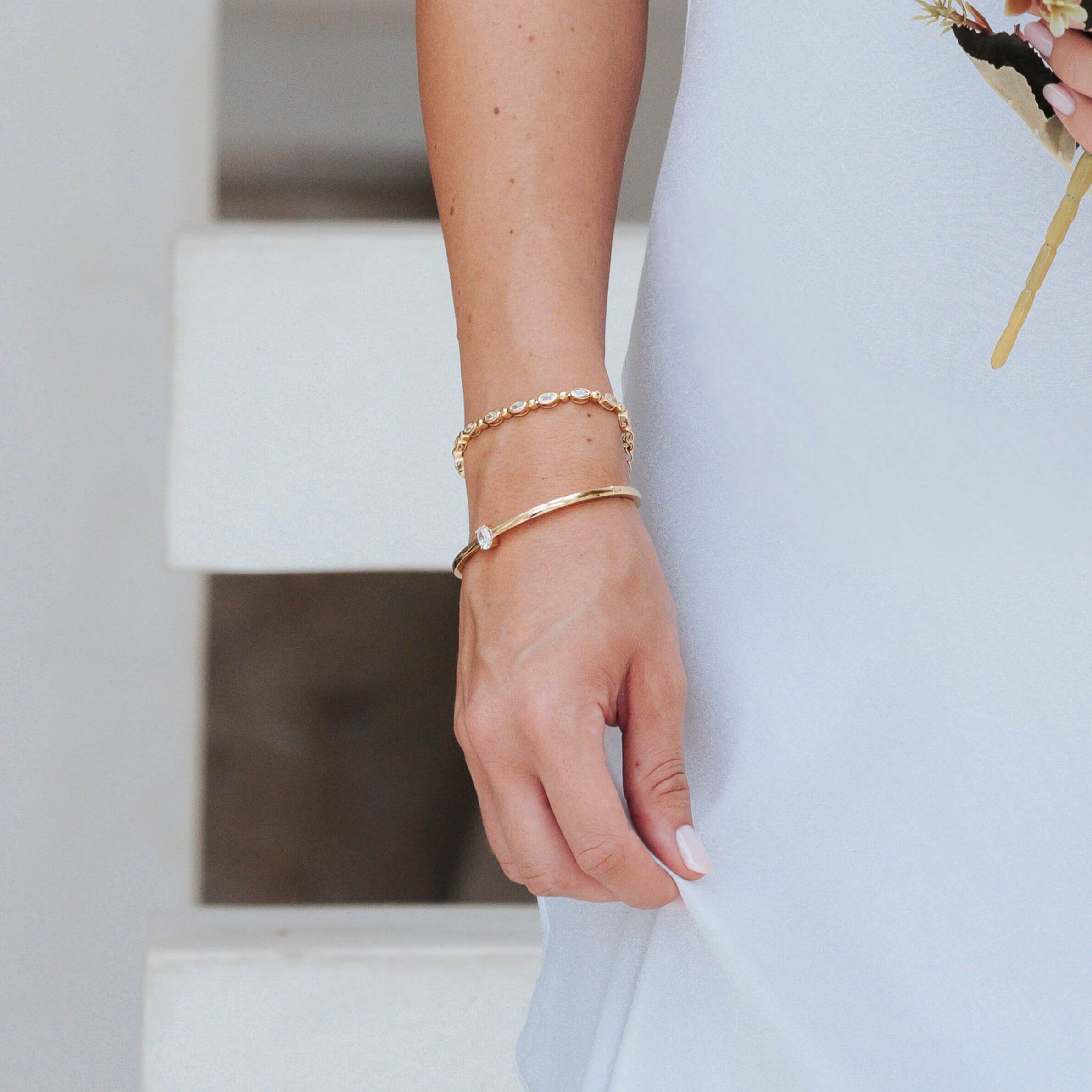 A close-up of a person's hand and wrist wearing two delicate gold bracelets, one beaded and one slim with a small clear stone, while wearing a white dress.