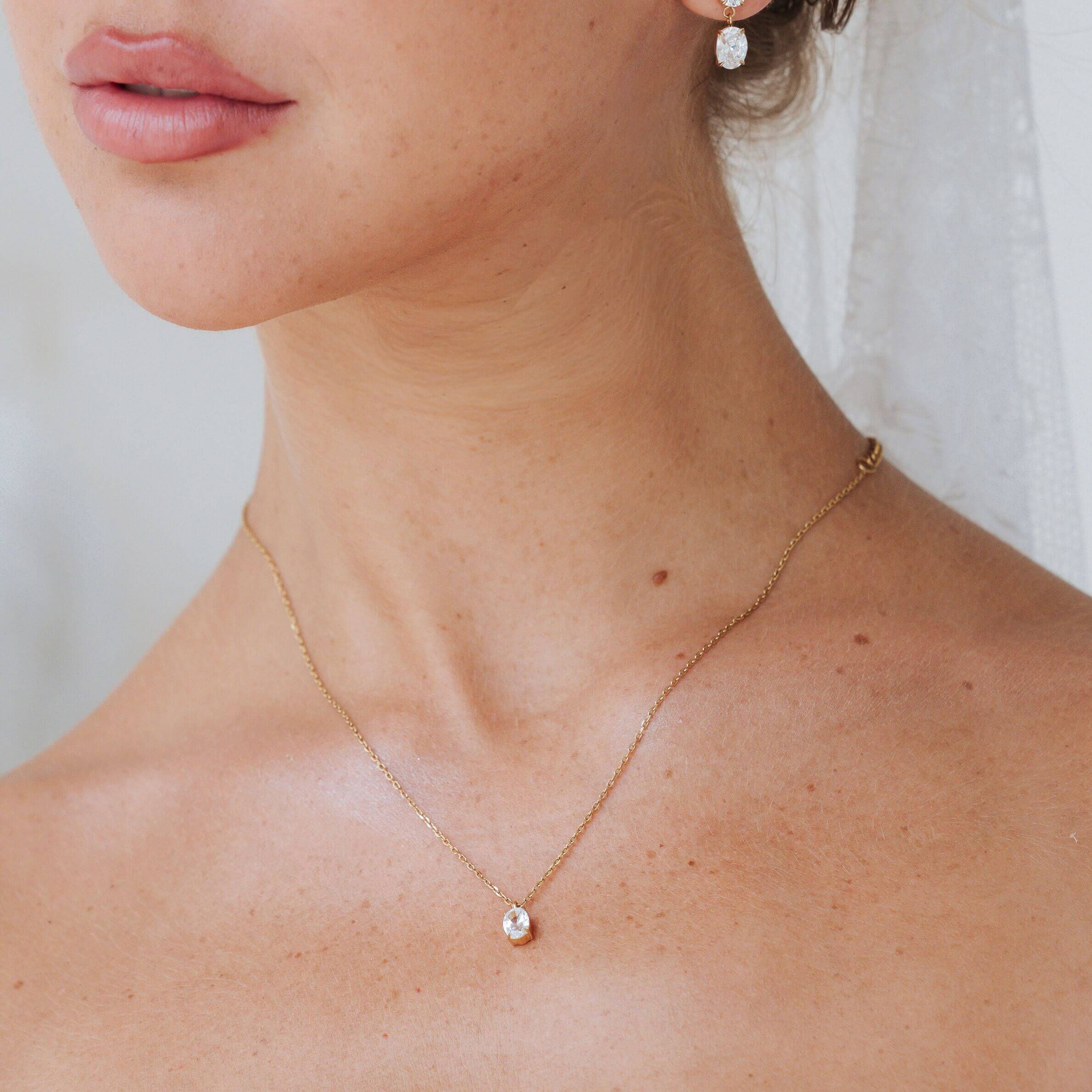 A close-up of a woman's neck and upper chest wearing a delicate gold chain necklace with a small round clear stone pendant, along with a matching drop earring.