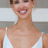 A close-up of a smiling bride wearing a delicate gold necklace with a small white diamond pendant, gold hoop earrings with pearl drops, and a white spaghetti-strap top.