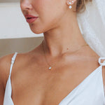 A close-up lifestyle shot of a woman wearing a delicate gold necklace with a small clear teardrop-shaped gemstone pendant, styled with a white bridal veil and white spaghetti-strap dress.