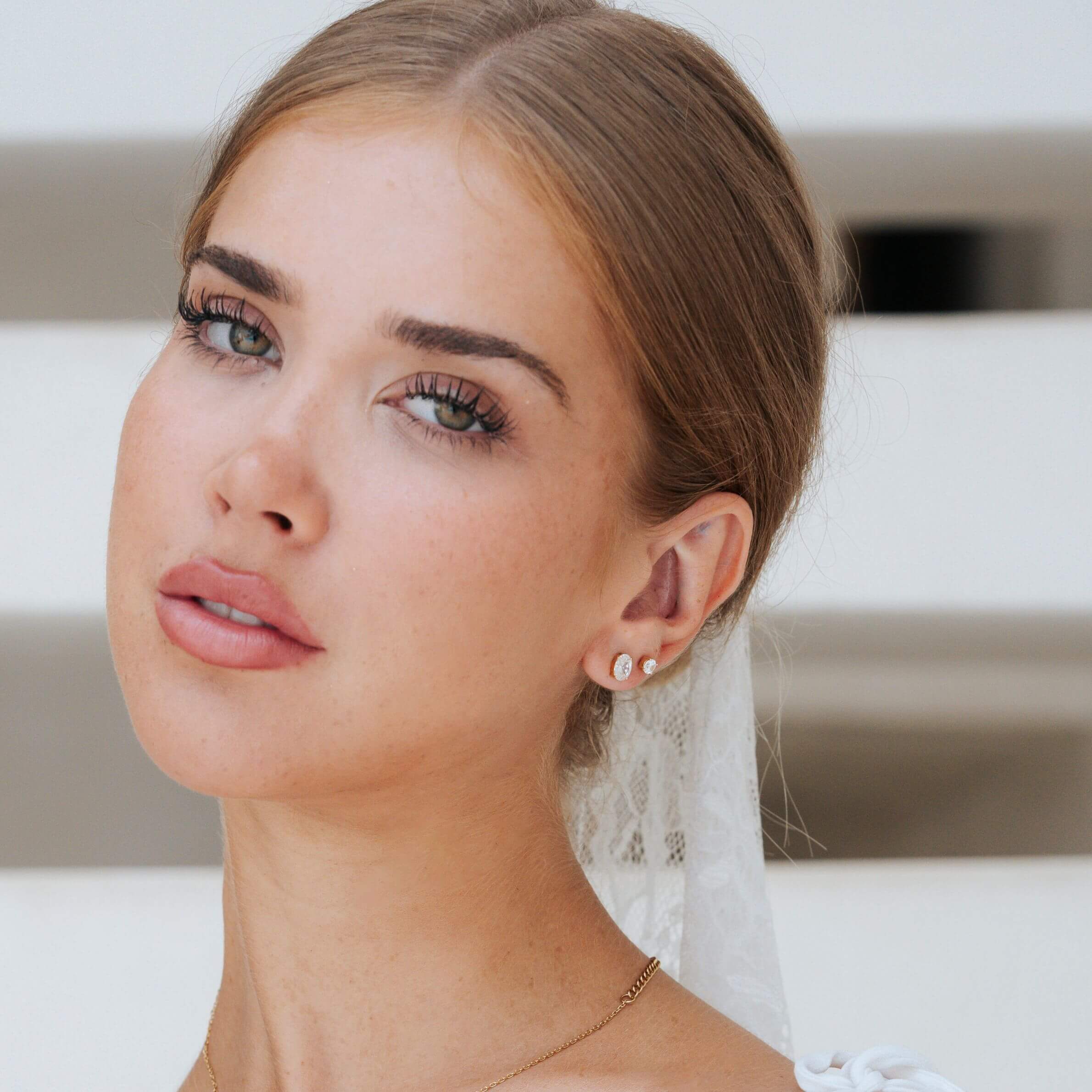A close-up portrait of a woman wearing small oval cut white zirconia stud earrings with gold-toned settings, styled with a white lace veil and delicate necklace against a soft white background.
