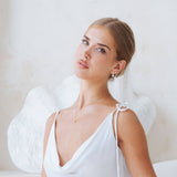 A woman with her hair pulled back wears a white spaghetti-strap wedding dress and delicate gold jewellery, including a necklace and earrings, posed in front of a soft white textured background with angel wings behind her.