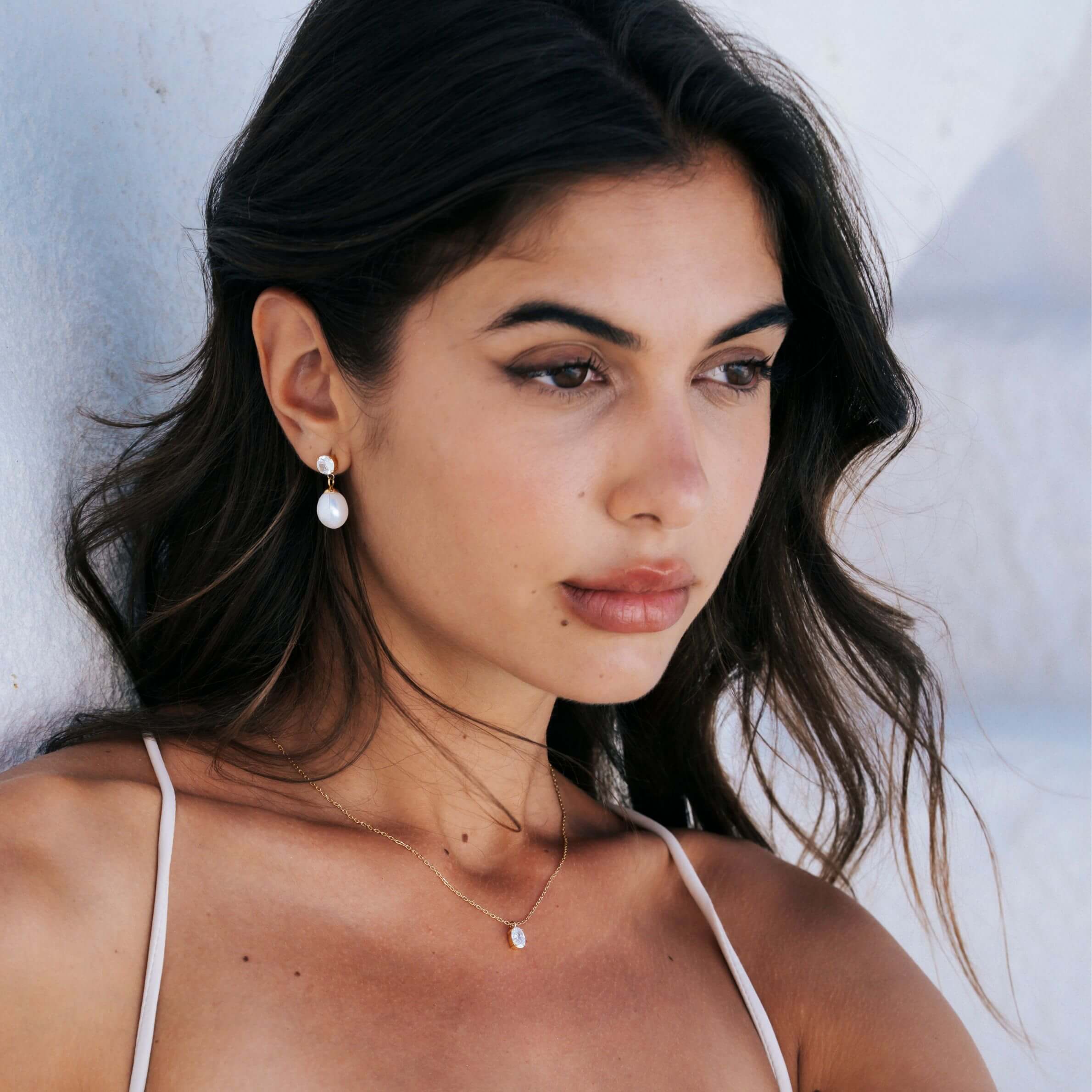 A close-up lifestyle shot of a woman with dark hair wearing a small white diamond and pearl drop earring and a delicate gold necklace with a small white stone, set against a light, textured wall. The jewellery the woman is wearing is designed by Mettle & Bloom for brides. 
