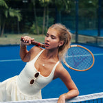 A woman on a blue tennis court holding a vintage tennis racket, wearing a cream-colored sleeveless dress and gold jewelry.