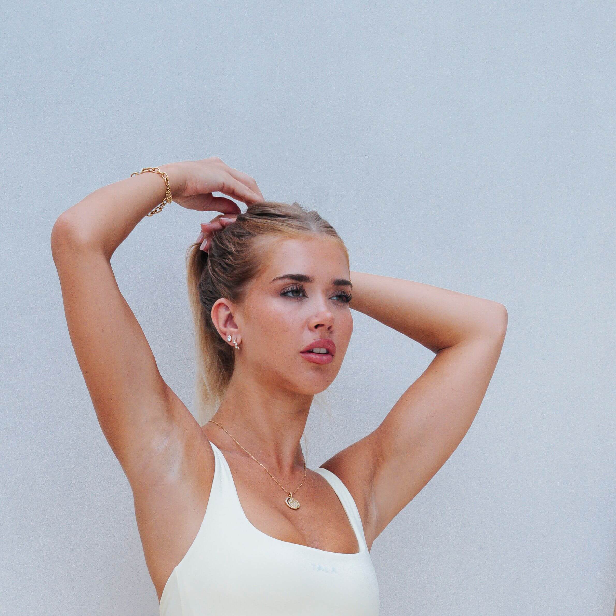 A woman with blonde hair tied back in a ponytail, wearing a white tank top and gold jewelry, posing against a light gray background.