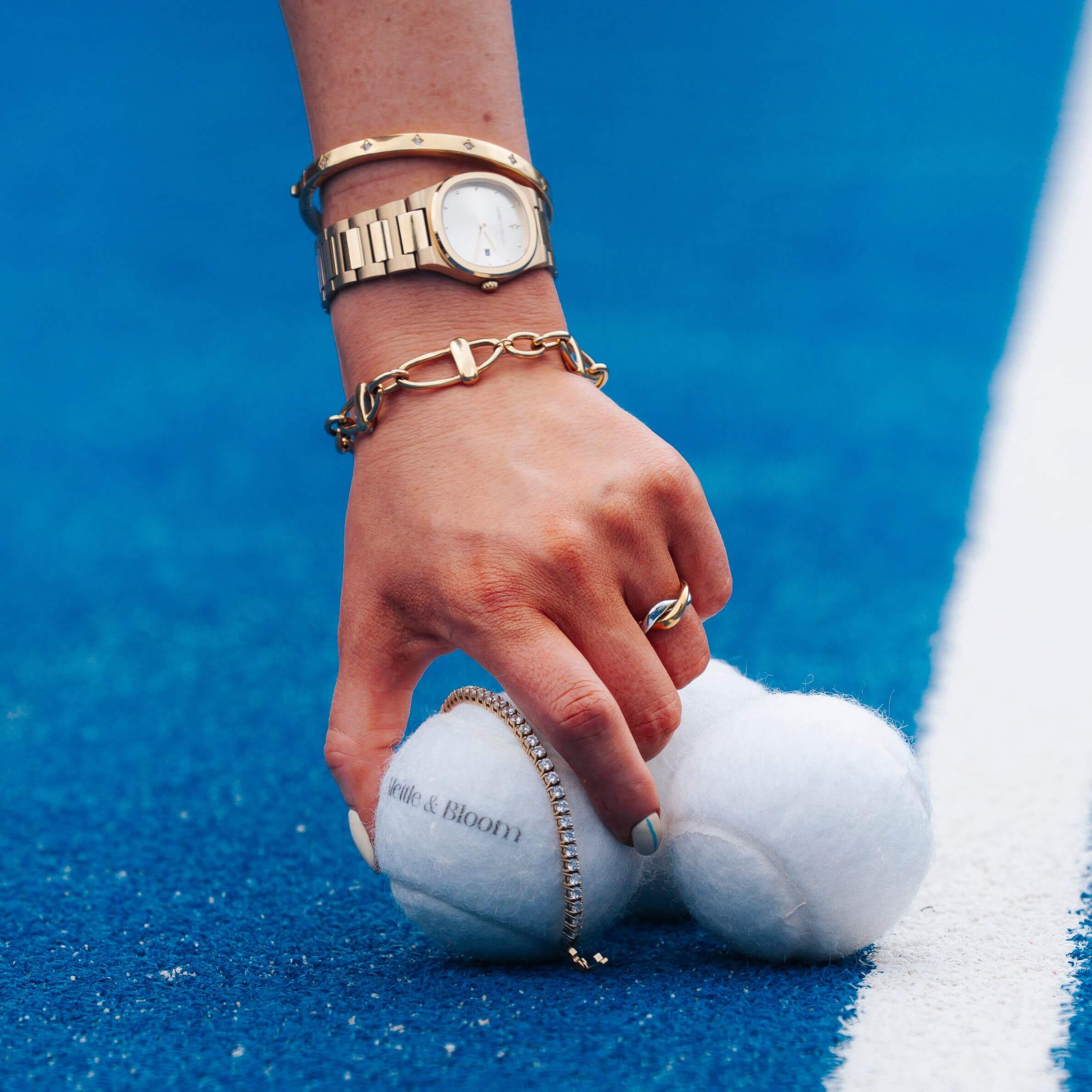 A close-up of a person's hand resting on two white tennis balls on a blue running track, wearing gold jewelry including a watch, bracelets, and rings.