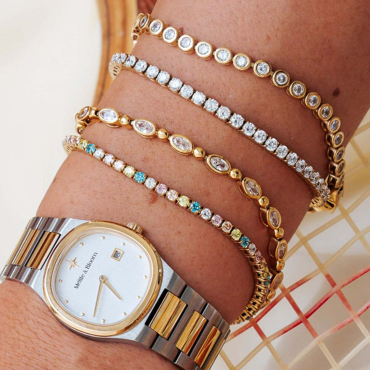 A close-up of a person's wrist wearing a two-tone stainless steel and gold watch, layered with four delicate gold bracelets adorned with clear crystals and small colored stones, set against a warm beige woven background.