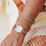 A close-up of a person's wrist wearing a two-tone stainless steel and gold watch, layered with four delicate gold bracelets adorned with clear crystals and small colored stones, set against a warm beige woven background.