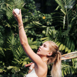 A woman in a white tennis dress prepares to serve, holding a white tennis ball overhead with her arm raised. She is wearing multiple gold bracelets and earrings, with a tennis racket visible behind her shoulder, set against a lush background of green tropical palm leaves.
