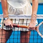 A close-up lifestyle shot of a tennis player at the net, wearing a cream-colored tennis dress and holding a Wilson racket. The player is accessorized with a silver watch, a gold bracelet, and stacked silver and rose gold bracelets on both wrists.