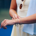 A close-up lifestyle shot of a woman wearing delicate gold and silver chain bracelets on her wrist, with her other hand resting nearby. She is wearing a cream-colored dress and a light blue towel is draped over her shoulder.