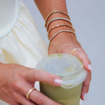 A close-up of a person's hands and wrist adorned with multiple delicate gold bracelets, including beaded chains and oval-link designs, while holding a clear-lidded cup.