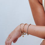 A close-up of a person's wrist wearing three delicate gold bracelets, including a tennis bracelet with clear stones and a small dangling charm, against a light gray background.