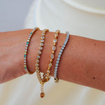 Four delicate bracelets stacked on a person's wrist, featuring gold and silver-toned metals with clear stones and a touch of turquoise on one bracelet, set against a soft white background.