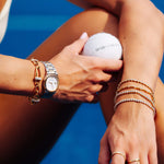 A close-up lifestyle shot of a woman's hands and arms adorned with multiple bracelets and rings, holding a white tennis ball against a vibrant blue background.