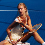 A woman in a cream tennis dress sits on a blue tennis court holding a tennis ball and a wooden racket.