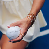 A close-up lifestyle shot of a person's hand holding a white tennis ball printed with the brand name 'Mettle & Bloom'. The person is wearing multiple gold bracelets and a gold ring, and is dressed in a cream-colored tennis dress against a blue tennis court background.