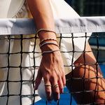 A close-up lifestyle shot of a person wearing multiple delicate tennis bracelets on their wrist and a matching tennis ring on their finger, sitting behind a tennis net on a blue court.