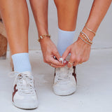 A close-up lifestyle shot of a person tying the laces of their white sneakers, wearing light blue crew socks and gold bracelets.