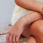 A close-up of a person's hands and arms wearing multiple delicate gold bracelets, including one with clear stones, on a rustic wooden surface.