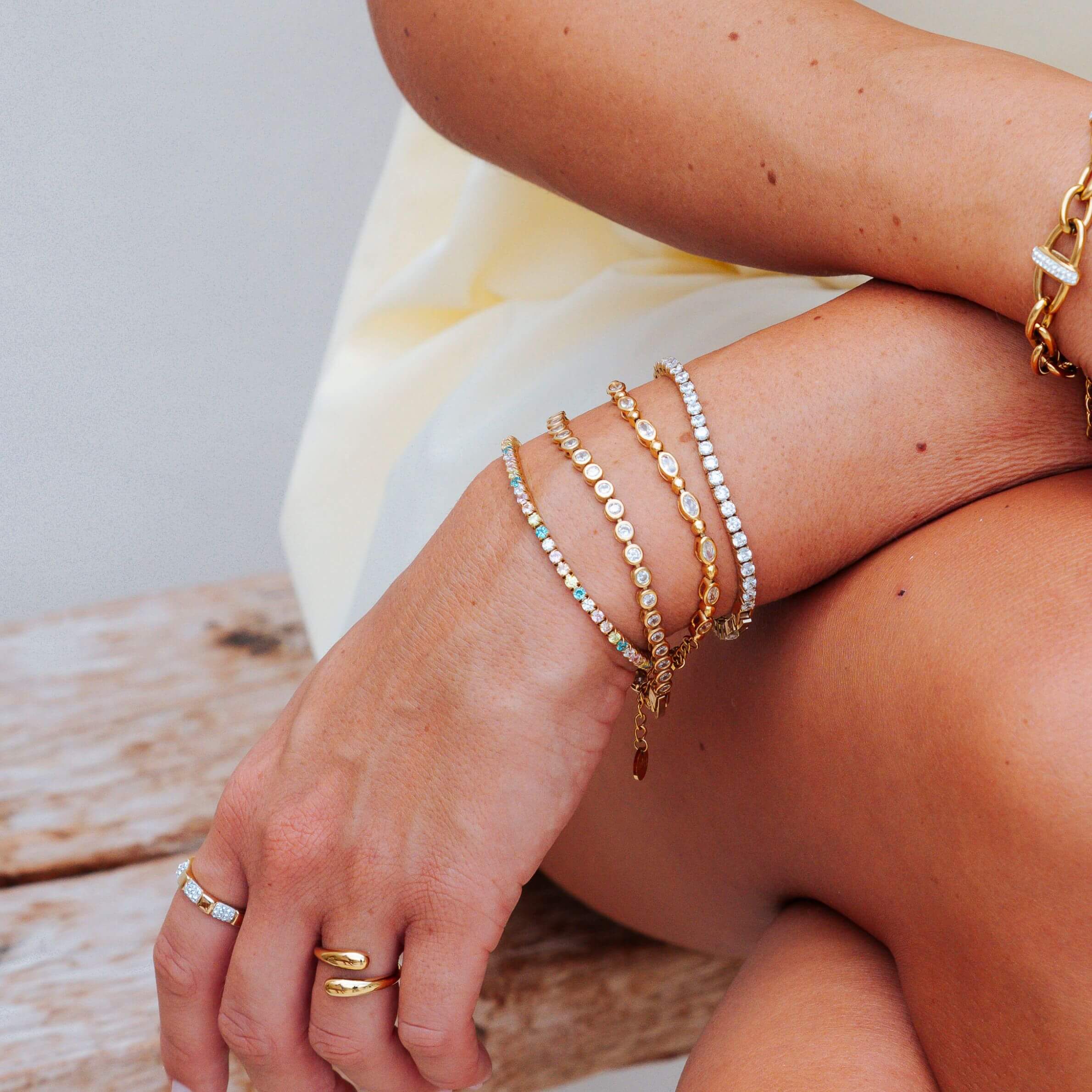 A close-up of a person's hands and arms wearing multiple delicate gold bracelets, including one with clear stones, on a rustic wooden surface.