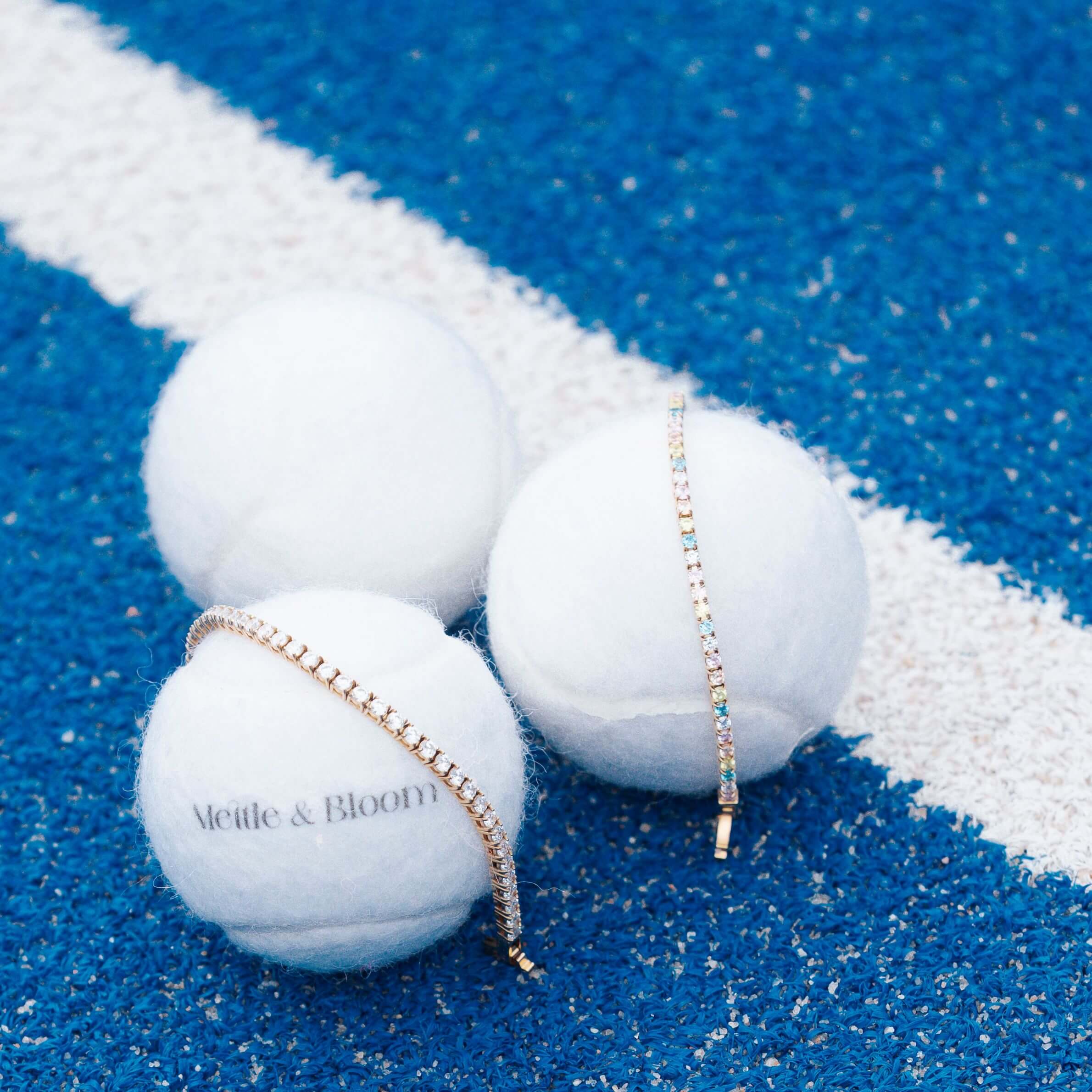 Three white tennis balls on a blue running track with a white line. Two tennis balls are white, and one is printed with the text 'Mettle & Bloom'.
