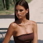 Woman wearing a strapless brown dress with layered necklaces outdoors. the sun is making her mettle and bloom necklaces glisten. 