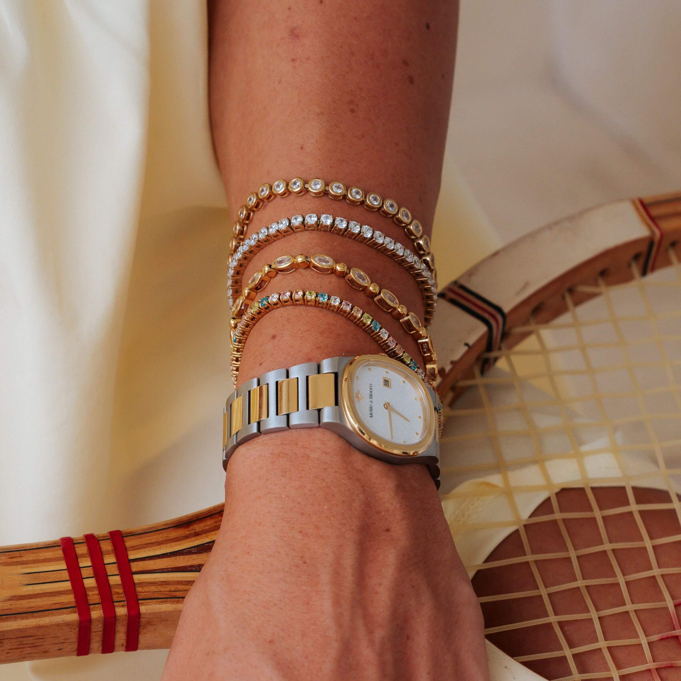 A close-up of a person's wrist wearing a gold and silver two-tone watch with stacked crystal bracelets, resting near a tennis racket on a light cream fabric background.