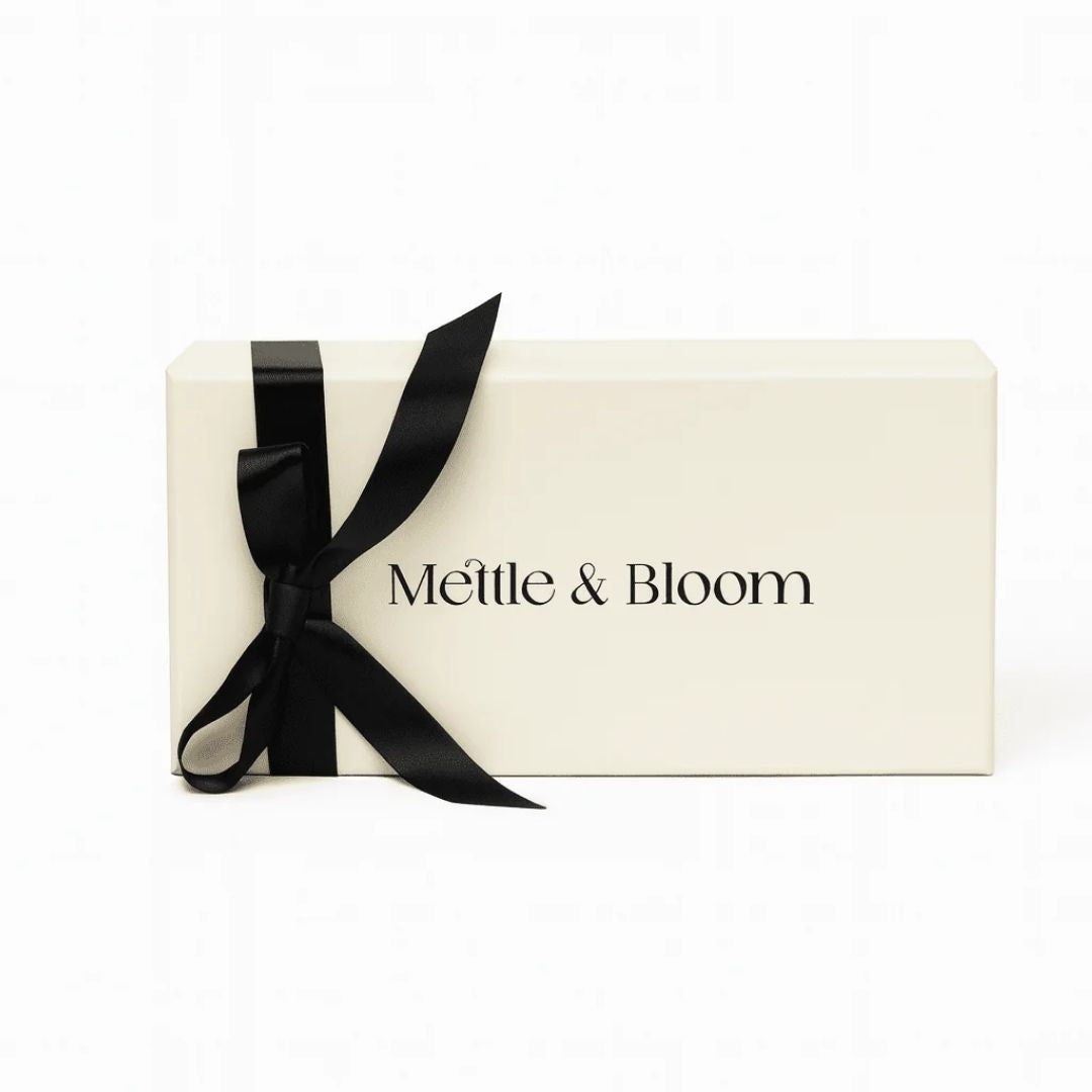 A cream-colored rectangular gift box with the brand name 'Mettle & Bloom' printed in black on the front, tied with a black satin ribbon bow, set against a plain white background.