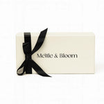 A cream-colored rectangular gift box with the brand name 'Mettle & Bloom' printed in black on the front, tied with a black satin ribbon bow, set against a plain white background.