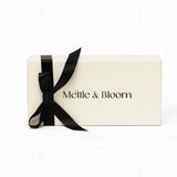 A cream-colored rectangular gift box with the brand name 'Mettle & Bloom' printed in black on the front, tied with a black satin ribbon bow, set against a plain white background.