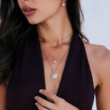 A close-up lifestyle shot of a model wearing layered gold necklaces, including a moon and star pendant, a matching star earring, and a gold ring, paired with a dark plum halter top.