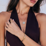 A close-up lifestyle shot of a model wearing a delicate gold chain necklace with a round, textured light star embellished pendant. The model is also wearing a gold ring and a gold bangle bracelet, and is wearing a dark plum halter top against a clean white background.